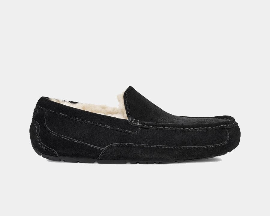A UGG Ascot suede slipper in black color with a rubber sole, featuring a sheepskin lining and a logo emboss.
